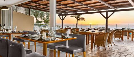 An outdoor dining area with tables and chairs, a wooden ceiling, and a view of the ocean at sunset.