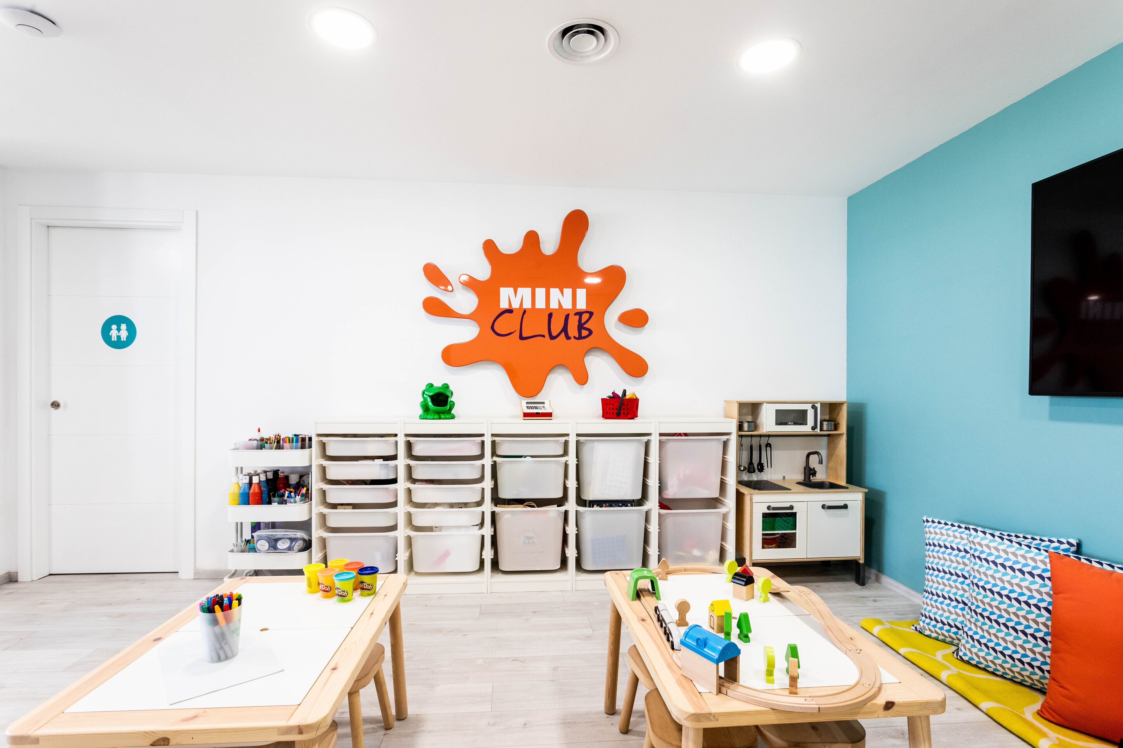 A children's playroom with two wooden tables, storage bins, and a 'Mini Club' sign on the wall.