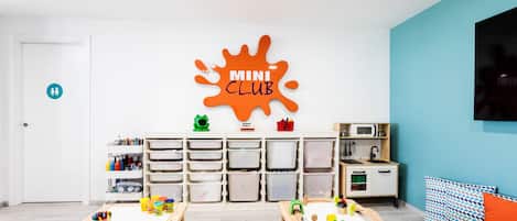 A children's playroom with two wooden tables, storage bins, and a 'Mini Club' sign on the wall.