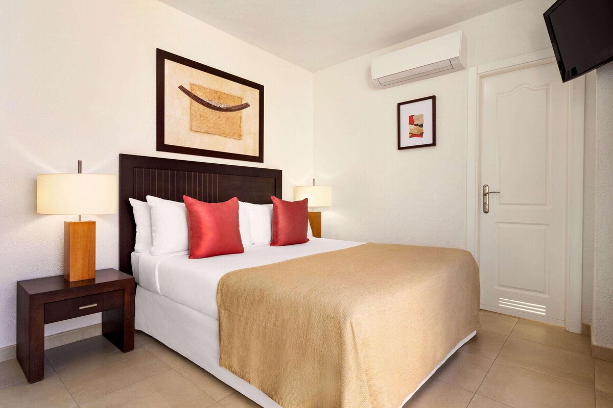 A hotel room with a bed, two bedside tables with lamps, a framed artwork above the bed, an air conditioning unit, a door, and a television mounted on the wall.