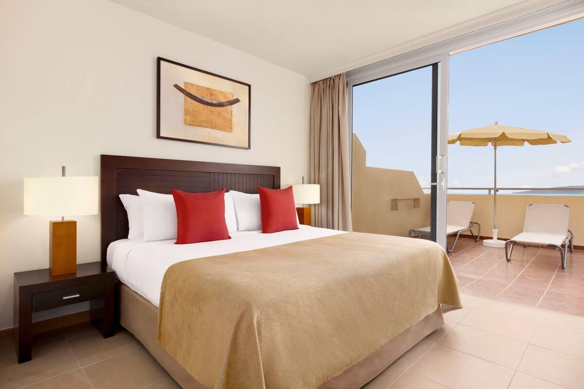 A hotel room with a large bed, two bedside tables with lamps, a framed artwork above the bed, a balcony with chairs and an umbrella, and a view of the sea.