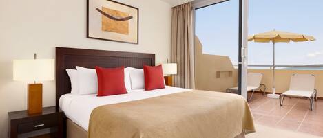 Habitación de hotel con una cama grande, dos mesitas de noche con lámparas, una obra de arte enmarcada sobre la cama, un balcón con sillas y una sombrilla, y vista al mar.