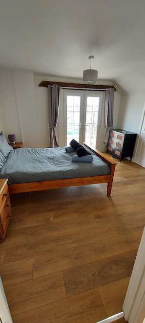 Un lit simple avec un cadre en bois et une literie grise, une commode en bois, un téléviseur posé sur un meuble et une fenêtre avec des rideaux.