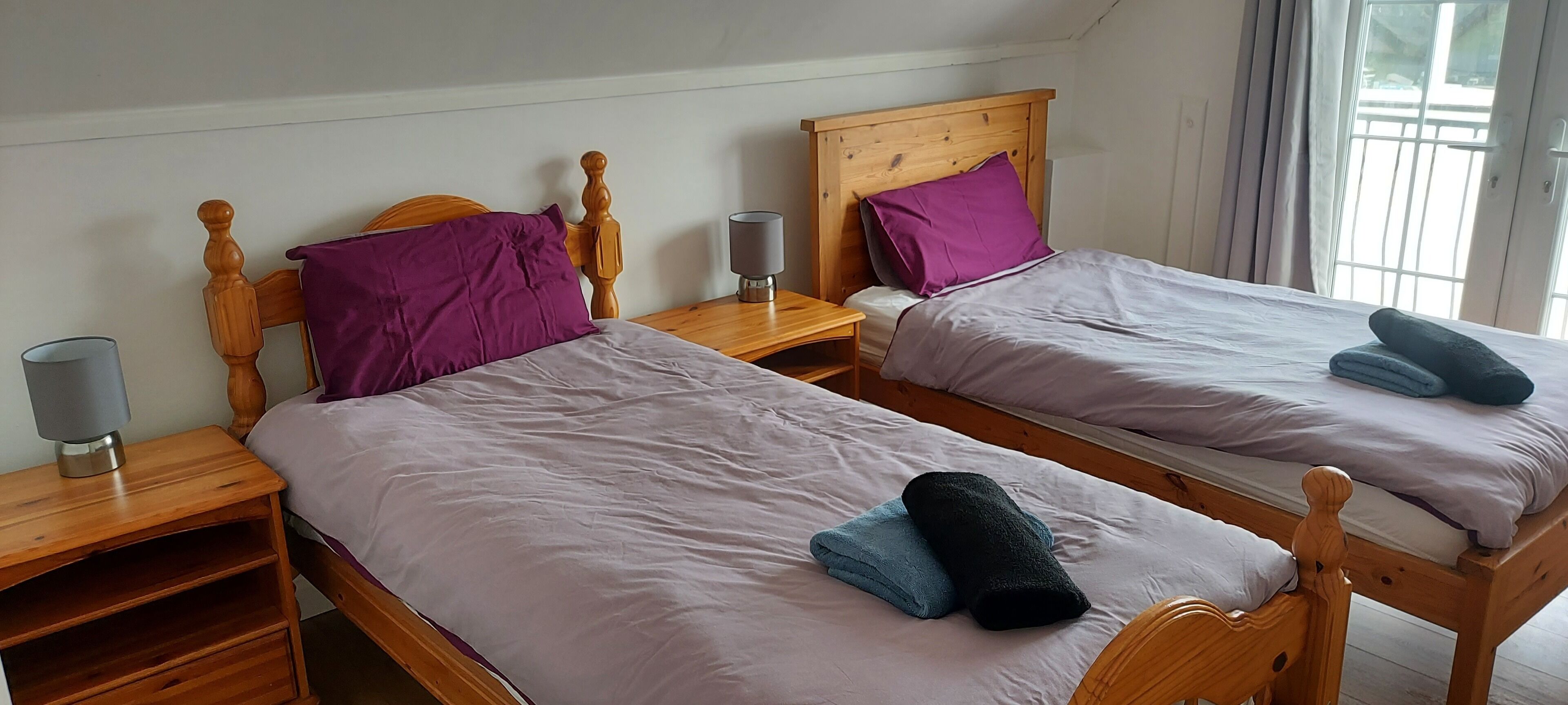 Two wooden beds with purple pillows and towels, a bedside table with a lamp, and a window with curtains.