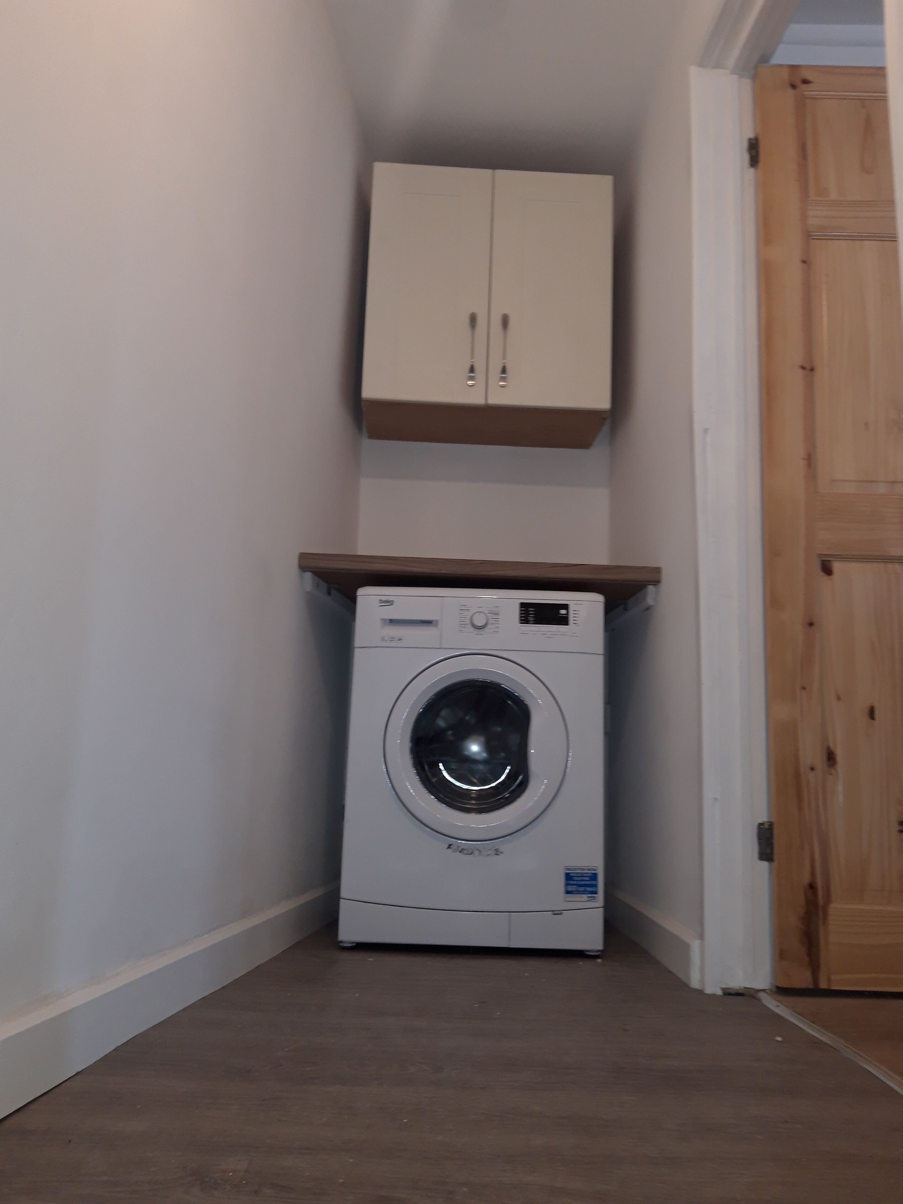 Une petite buanderie équipée d’un lave-linge à chargement frontal, d’un meuble en bois placé au-dessus, et d’une porte en bois située à droite.