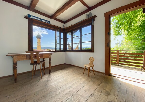 A room with a wooden desk, two chairs, a small wooden stool, and a large window with a view of a tower and greenery outside.