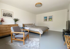 A modern bedroom with a bed, a wooden chair, a small table, a TV, and artwork on the walls.