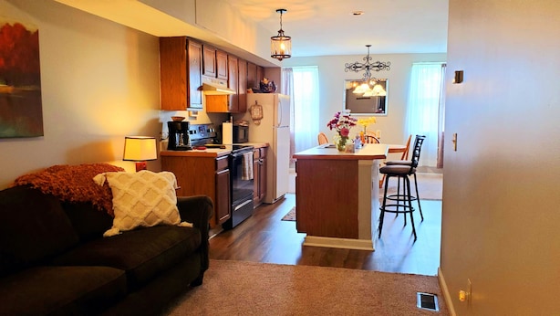 A cozy living room with a dark brown sofa, a coffee maker, and a lamp. The kitchen features wooden cabinets, a refrigerator, and a dining area with a chair and table.
