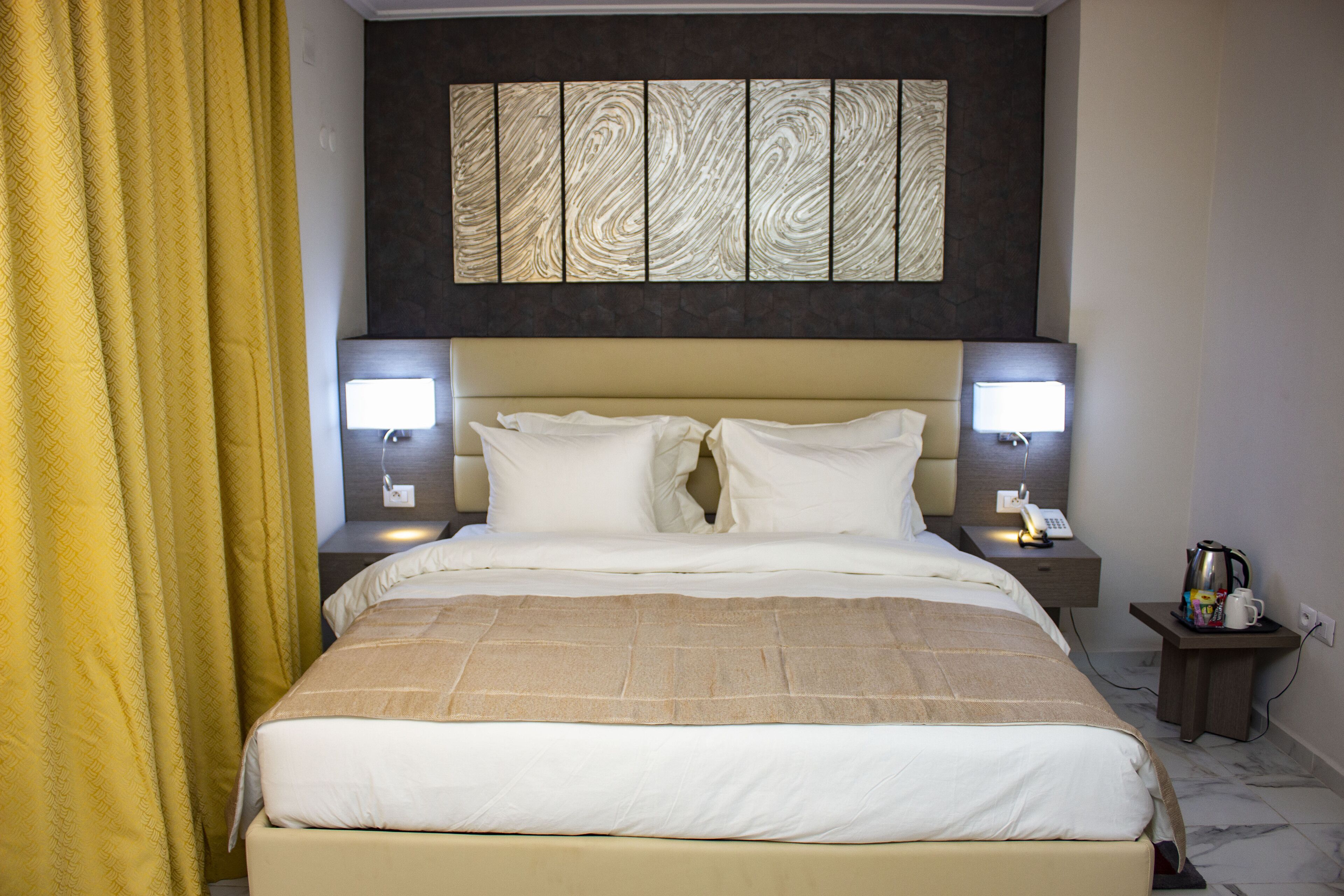 A hotel room with a large bed, two bedside lamps, a nightstand with a coffee maker, and a wall art piece above the bed.