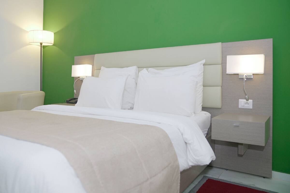 A hotel room with a bed, two bedside tables, a nightstand, a lamp, and a green wall.
