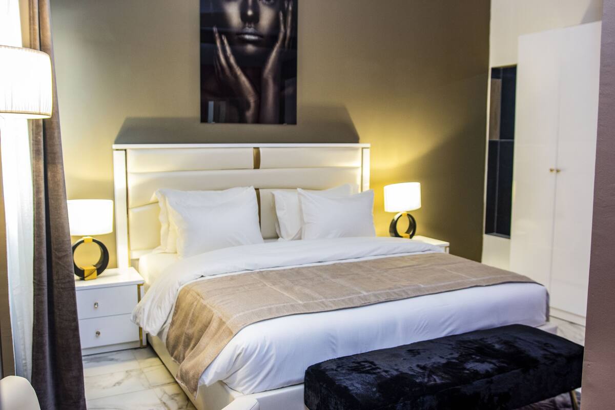 A hotel room with a large bed, bedside tables, a nightstand, a black ottoman, and a wall-mounted artwork.