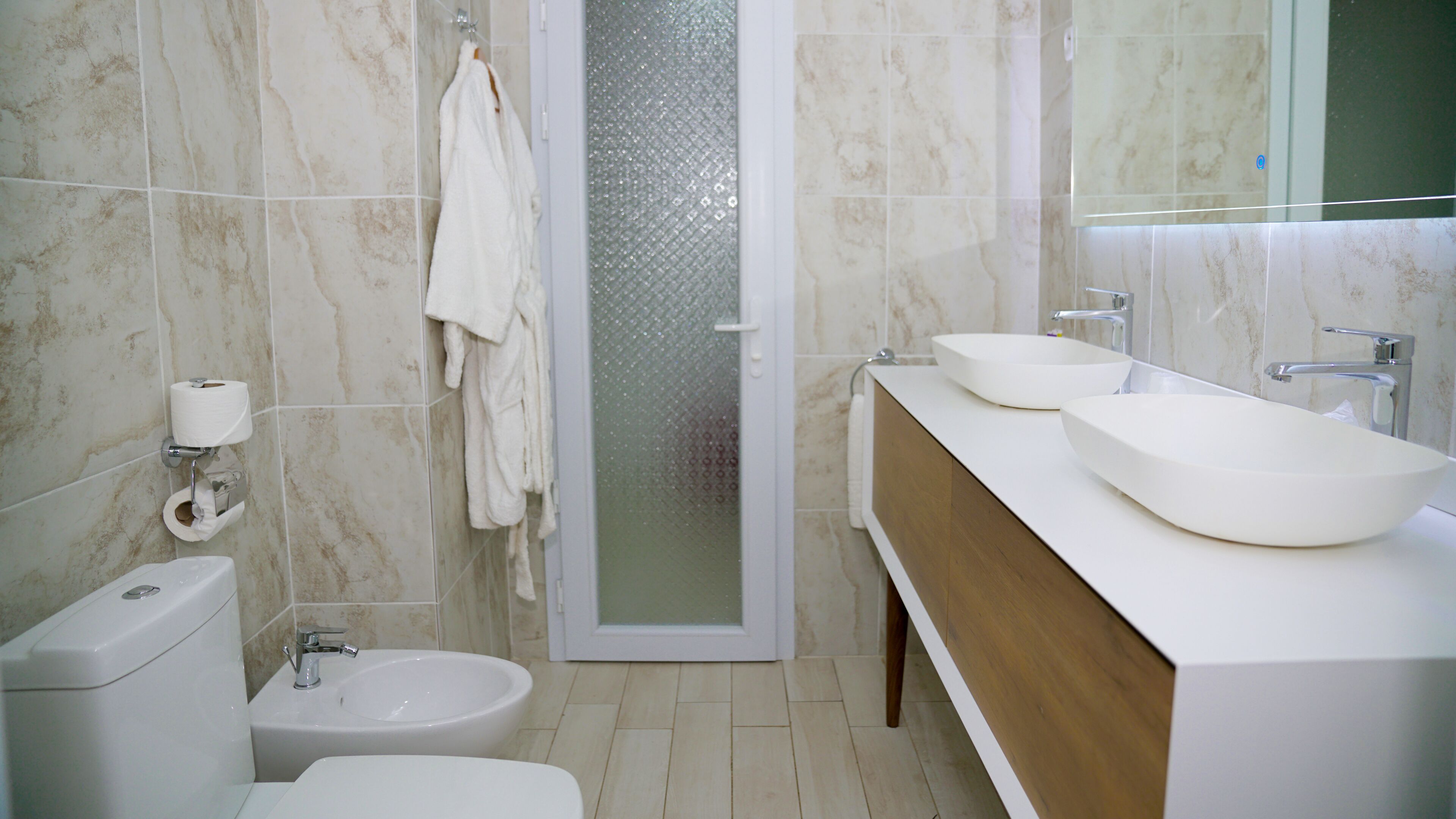 A modern bathroom with a toilet, bidet, two sinks, and a shower area.
