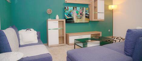 Una sala de estar moderna con una pared verde azulado, un televisor de pantalla plana, un mueble de madera, una mesa de centro de vidrio y dos sofás morados.