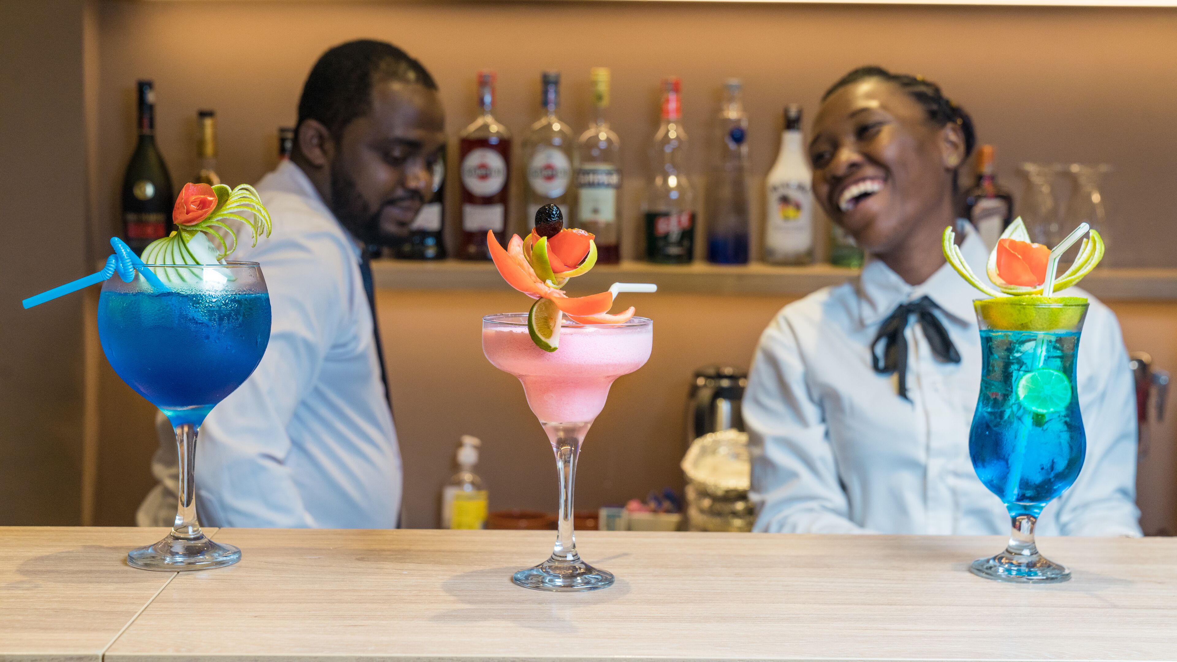 Three cocktails on a bar counter with a variety of bottles in the background.