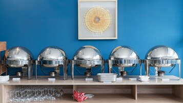 Una mesa de madera con cinco grandes fuentes de plata para buffet, una obra de arte enmarcada arriba y una pared azul de fondo.
