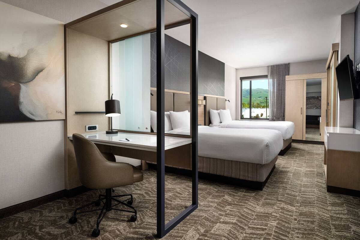 A hotel room with a desk, two beds, a large mirror, and a view of the outdoors.