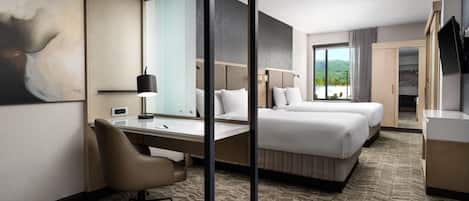 A hotel room with a desk, two beds, a large mirror, and a view of the outdoors.