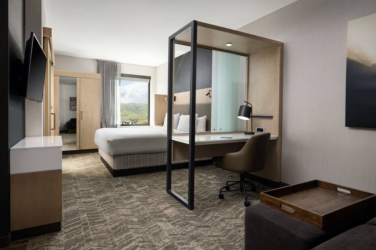 A hotel room with a bed, desk, chair, TV, and a large window with a view of hills.