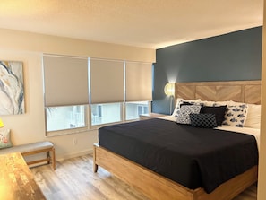 Ein Schlafzimmer mit einem Bett, einer Bank, einem Nachttisch, einer Lampe und einem Fenster mit Jalousien.