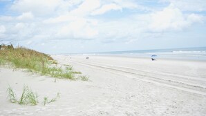 Une vaste plage de sable fin, avec des vagues douces, un ciel dégagé et une végétation clairsemée.