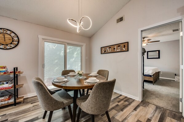 Un coin repas avec une table ronde dressée pour quatre personnes, entourée de chaises rembourrées. Une horloge en bois et un tableau encadré ornent les murs.