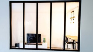 Una habitación con un ventanal grande dividido en cinco secciones, un televisor sobre un mueble bajo, una mesa con sillas y una lámpara colgante.