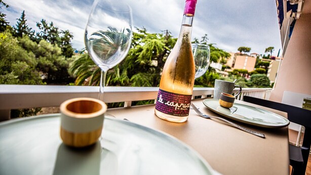 Una mesa con una botella de vino, dos copas de vino, un vaso de madera y un plato en un balcón con vistas a un jardín.