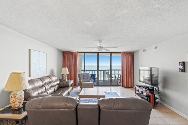 Ein Wohnzimmer mit einer Couch, Sesseln, einem Couchtisch, einem Fernseher und einem Balkon mit Meerblick.