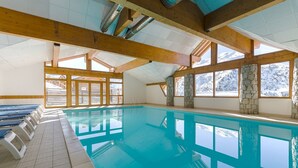 Une piscine intérieure à l’eau d’un bleu cristallin, agrémentée de poutres en bois, de piliers en pierre et de grandes fenêtres offrant une vue sur des montagnes enneigées.