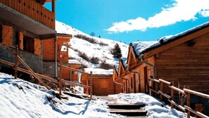 Une cabane en bois, avec des fondations en pierre, un escalier en bois menant à l’entrée, et une montagne enneigée en arrière-plan.