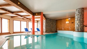 Una piscina interior con agua cristalina, vigas de madera, paredes de piedra y sillones de descanso azules.