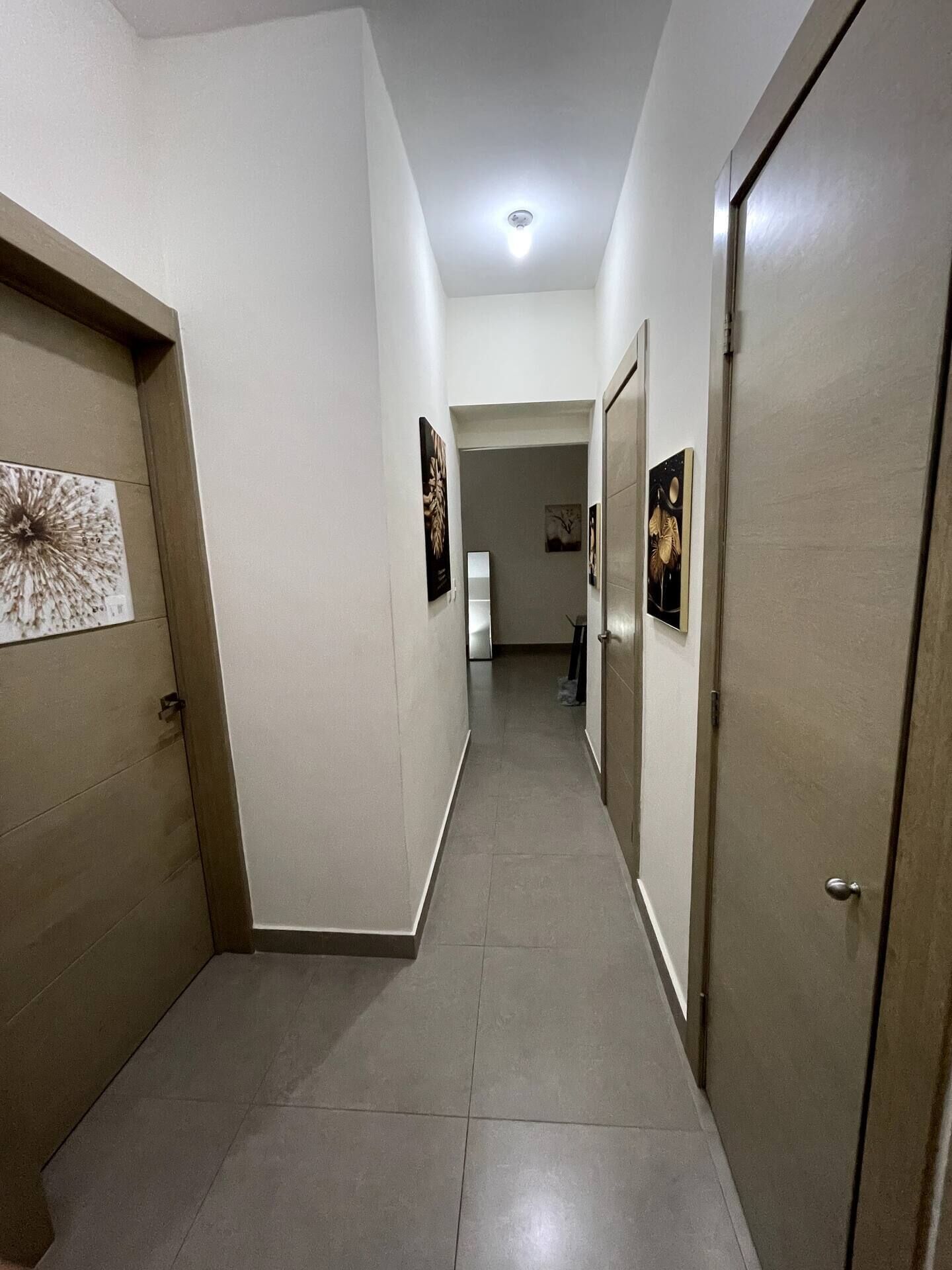 Um corredor com piso ladrilhado, paredes brancas e portas em ambos os lados. Quadros emoldurados adornam as paredes, e uma luminária ilumina o teto.