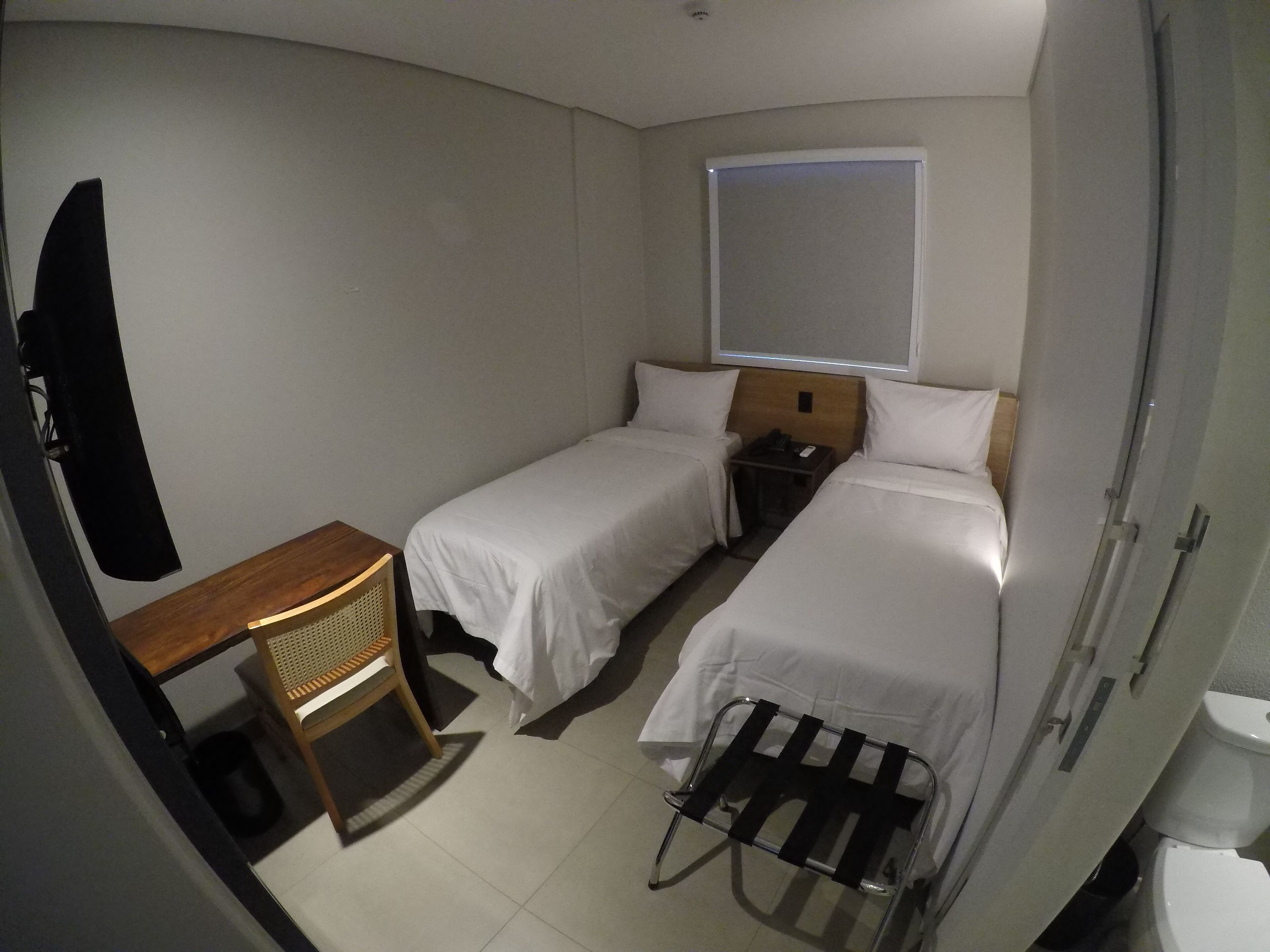 Um quarto de hotel pequeno com duas camas de solteiro, uma mesa de cabeceira de madeira, uma pequena mesa, uma cadeira e uma janela com persianas.