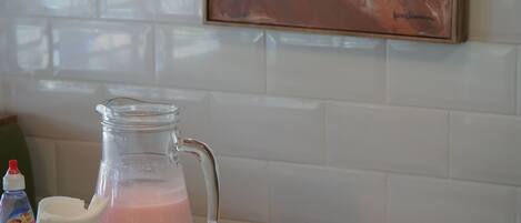 A breakfast table with various food items, a pitcher of green juice, a glass of pink smoothie, and a painting on the wall.