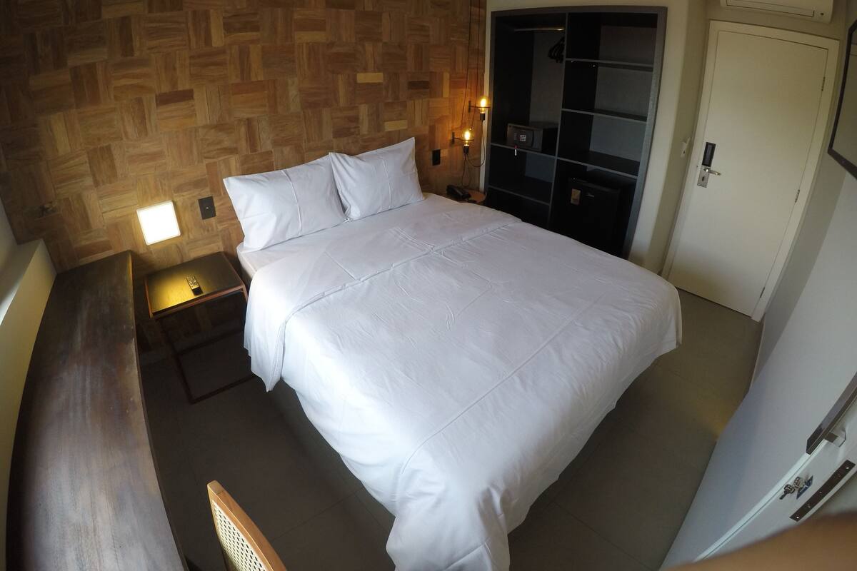 Un lit bien fait, avec du linge de lit blanc, une tête de lit en bois, une table de chevet avec une lampe et une armoire avec un miroir.