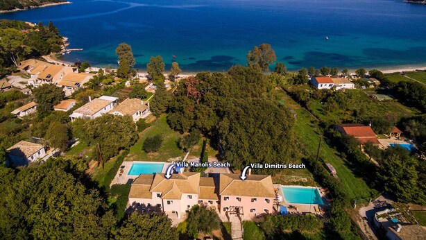 An aerial view of a coastal area with Villa Manolis Beach and Villa Dimitris Beach, featuring two swimming pools, lush greenery, and a backdrop of the sea and mountains.