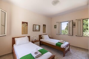 A hotel room with two single beds, a small bedside table with a lamp, a large window with multiple panes, and a ceiling light fixture.