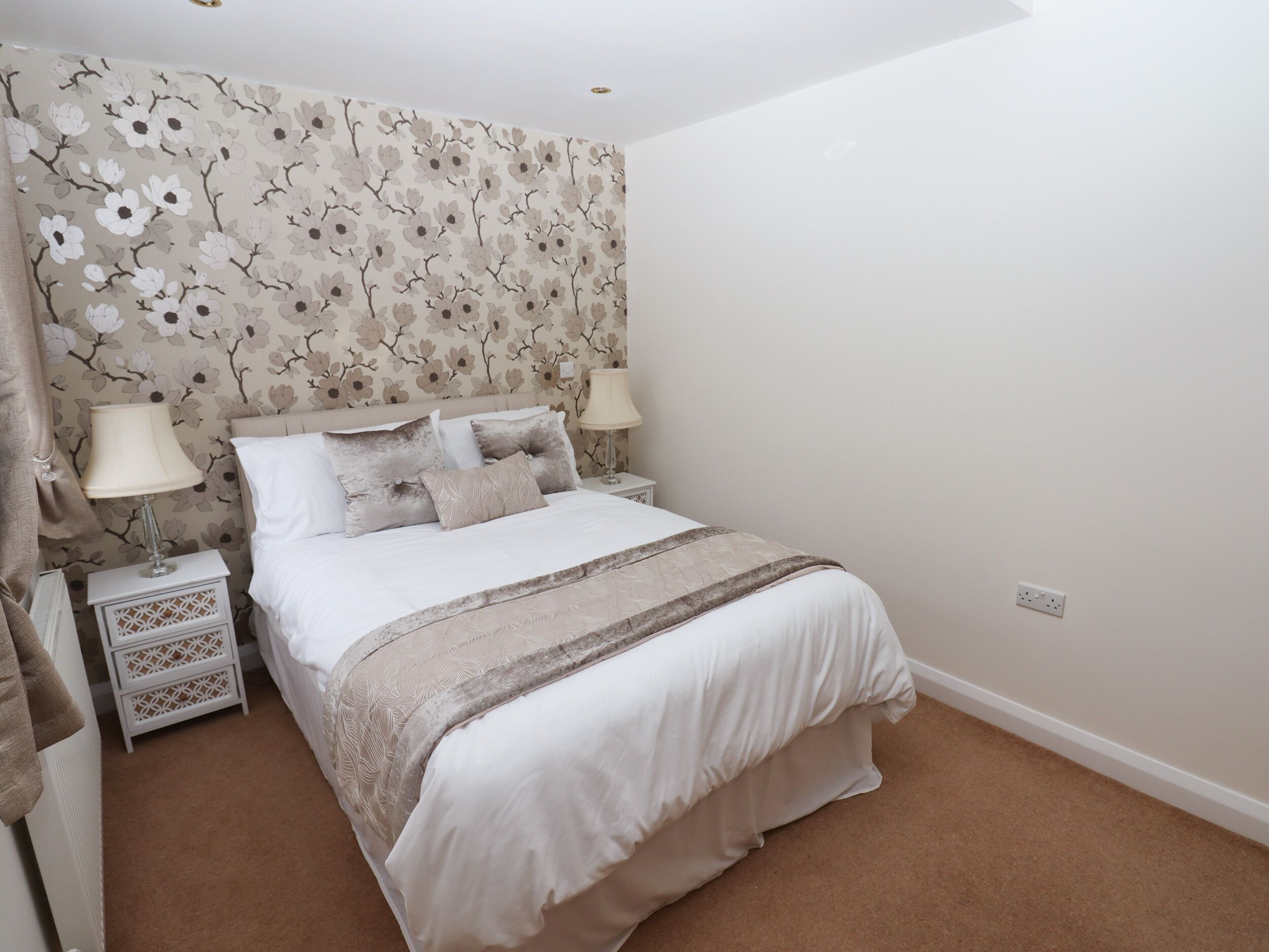 A neatly made bed with a floral wallpaper headboard, two bedside lamps, and a white nightstand with drawers.