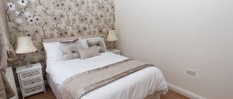 A neatly made bed with a floral wallpaper headboard, two bedside lamps, and a white nightstand with drawers.