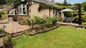 A stone house with a tiled patio, a garden with various plants, and a dining area with outdoor furniture. - Woodlands (Matlock)