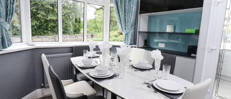 A dining area with a table set for four, white chairs, and a view of greenery through large windows.