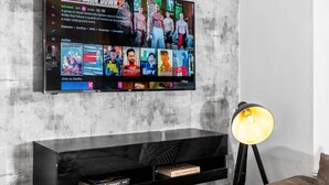 Un televisore montato su una parete in cemento mostra un film di combattimento. Sotto la TV, un mobile porta TV nero con una lampada e dei libri.