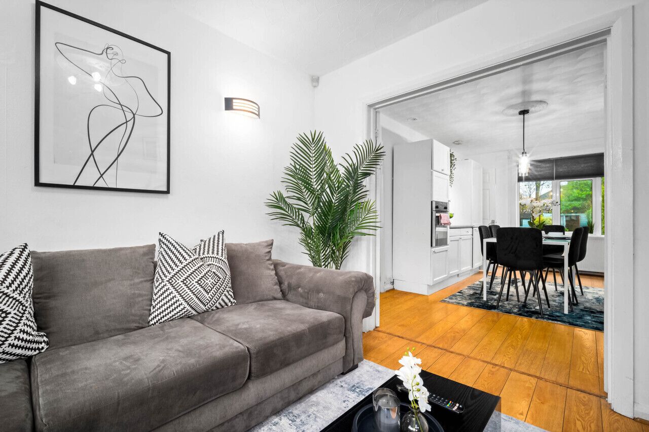 Una sala de estar moderna con un sofá gris, almohadones estampados en blanco y negro, un cuadro abstracto grande, una planta en maceta y una zona de comedor con una mesa y sillas negras.
