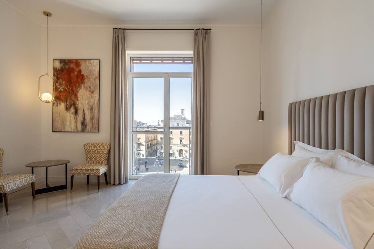 Habitación de hotel con una cama grande, vista de la ciudad por la ventana, un cuadro en la pared y una pequeña mesa con sillas.