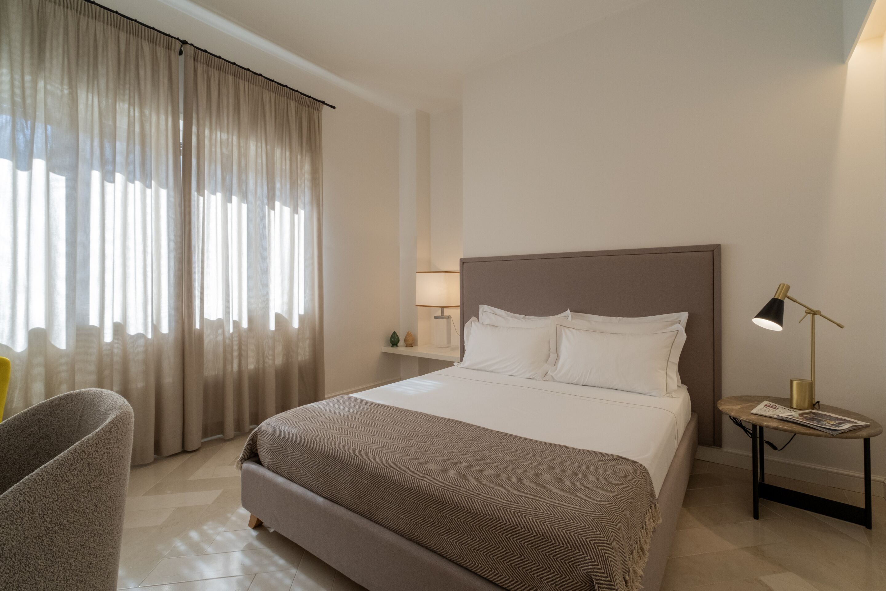 Una habitación de hotel con una cama grande, una mesita de noche con lámpara, una silla y una ventana con cortinas.