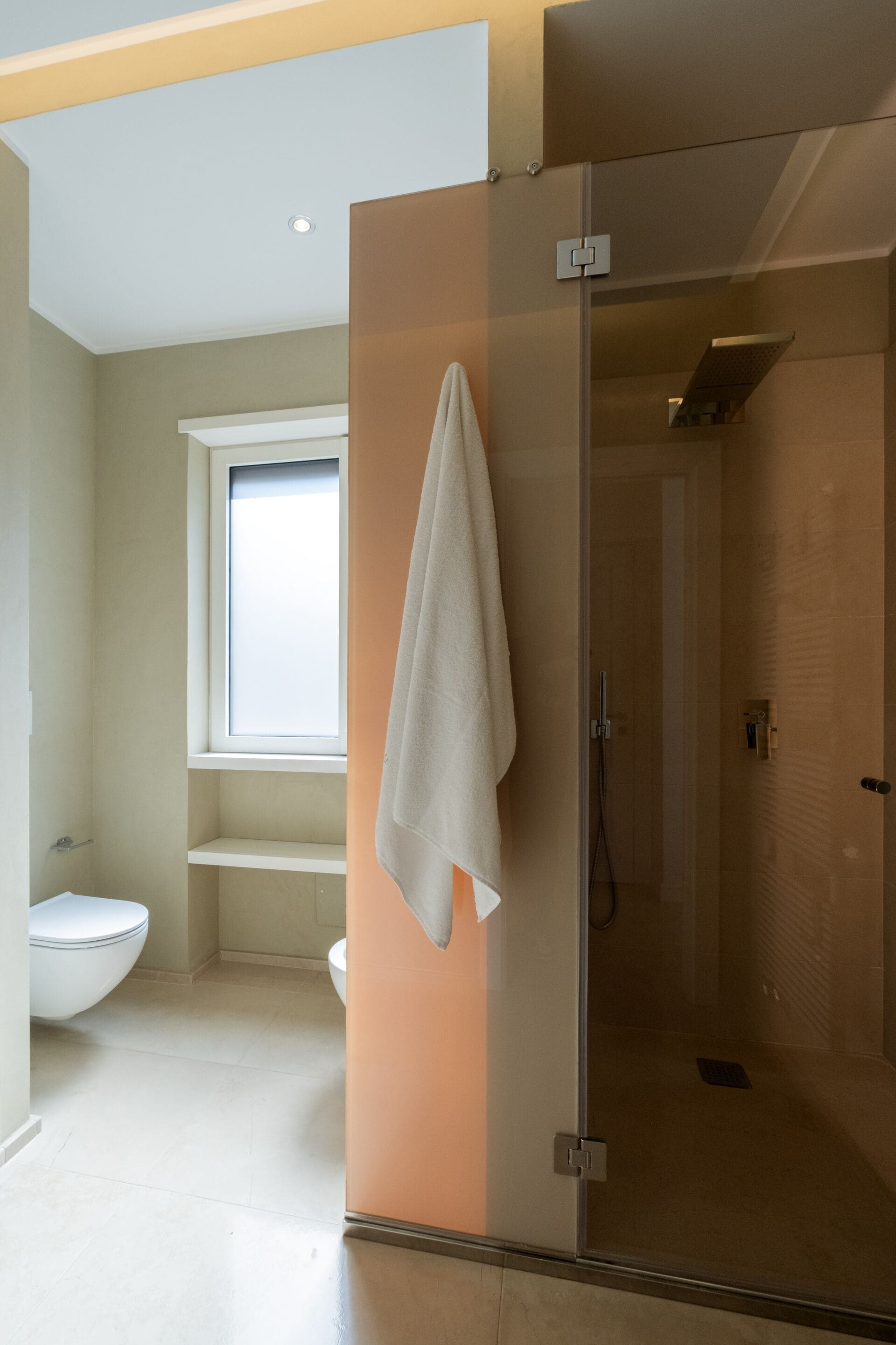 A modern bathroom with a glass shower enclosure, a toilet, and a window above the sink.