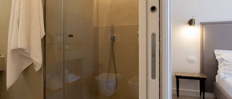 A modern bathroom with a glass-enclosed shower, a white towel hanging on the wall, and a bedside table with a lamp.