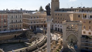 An ancient amphitheater surrounded by classical architecture, with a statue and a modern kiosk nearby. - Abalto Suites & Rooms (Lecce)