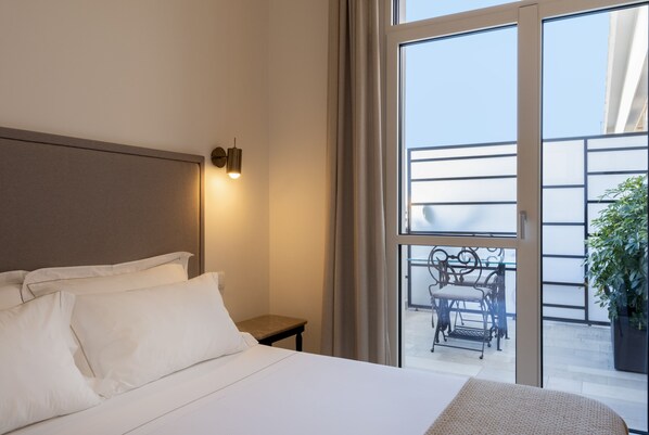 Habitación de hotel con una cama grande, una mesita de noche con lámpara y vistas al balcón con mobiliario exterior.