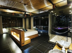 A modern hotel room with a wooden bed, a glass-enclosed bathroom, and a view of the outdoors. - Kura Design Villas (Ballena)